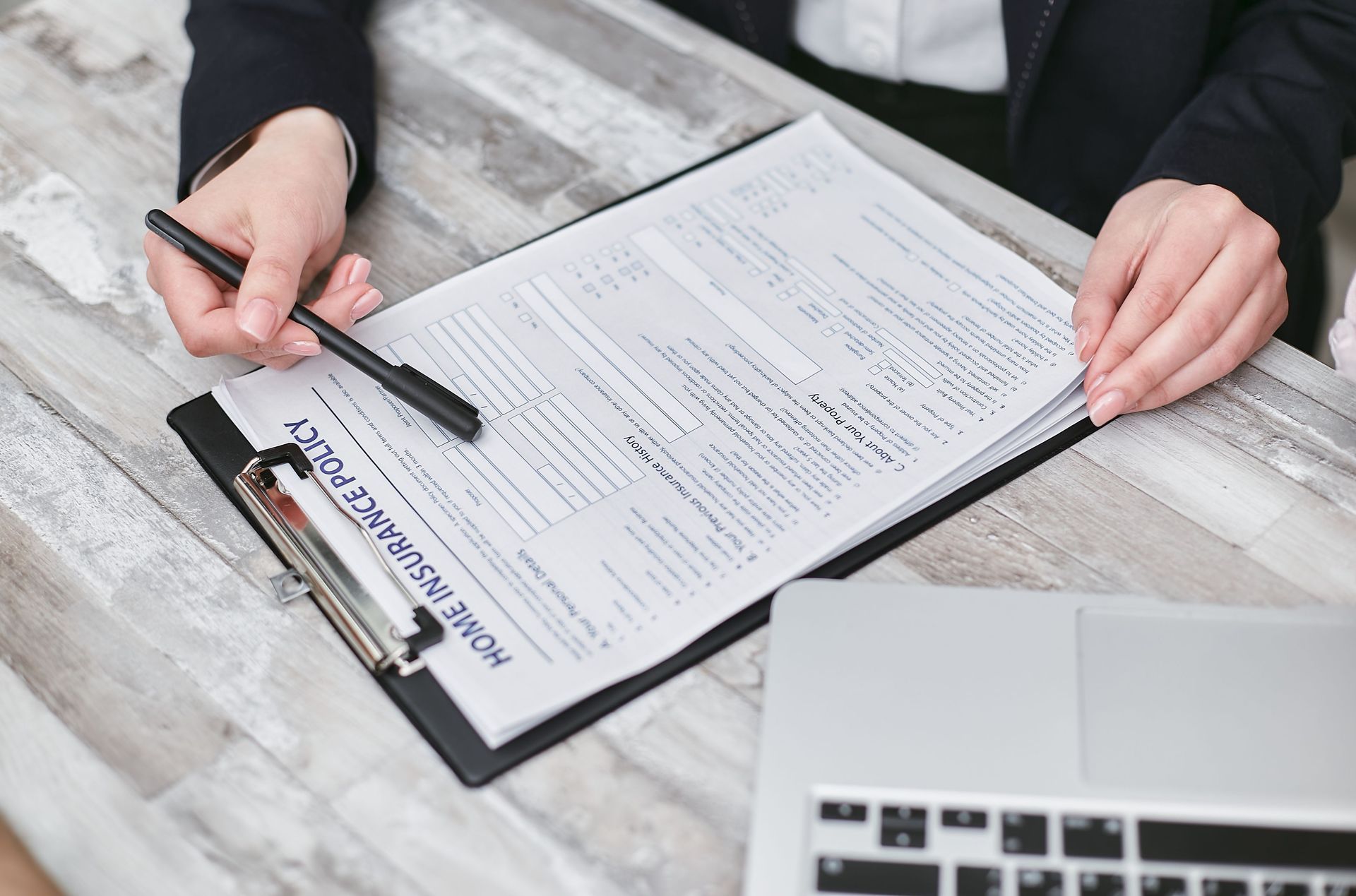 Hands writing on a clipboard with a pen beside a laptop on a light wooden desk