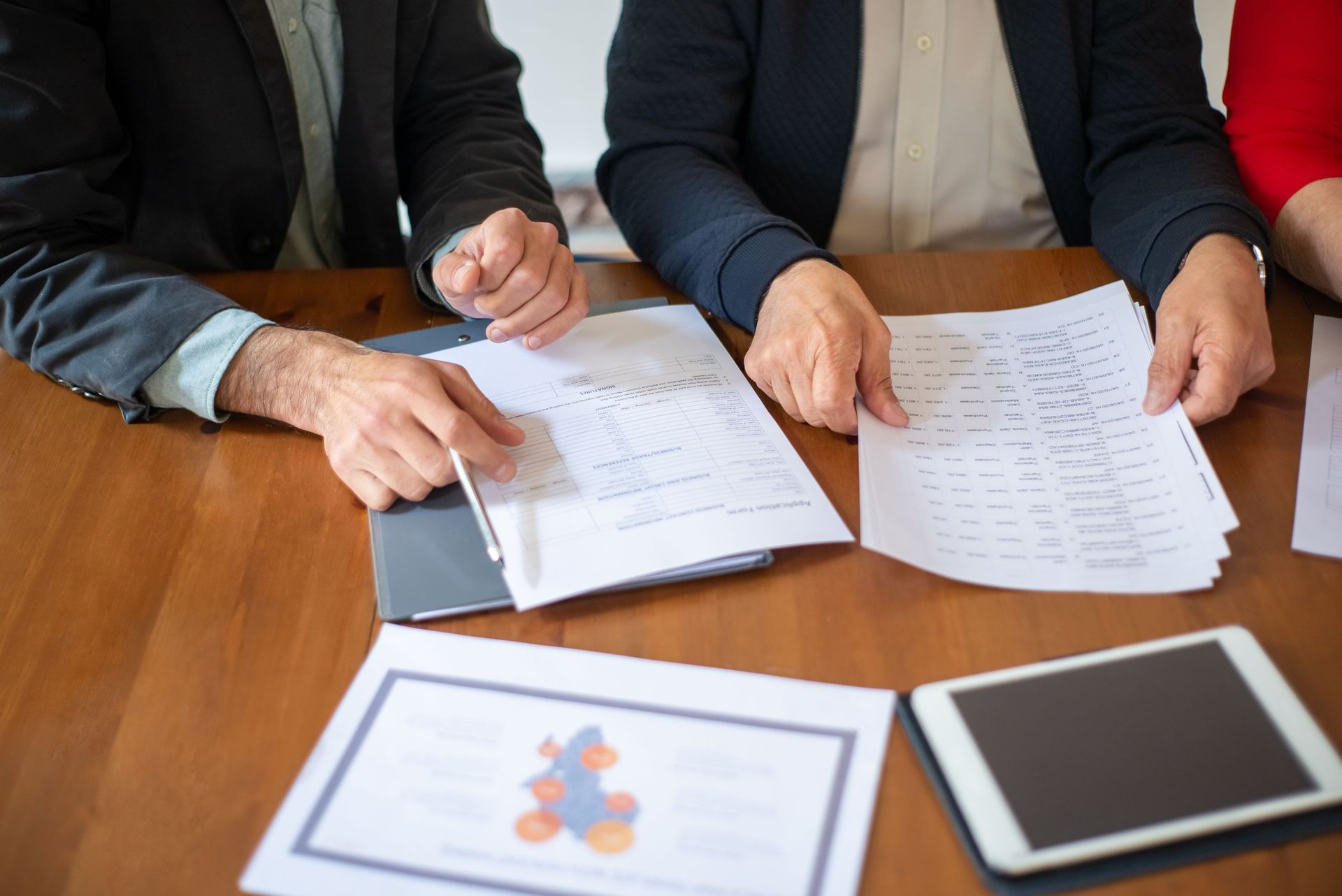 Three people in business attire review documents at a wooden conference table with a tablet and chart.