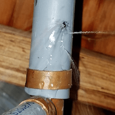 Gray pipe with a hole spurting water, connected to copper piping in a wooden setting.