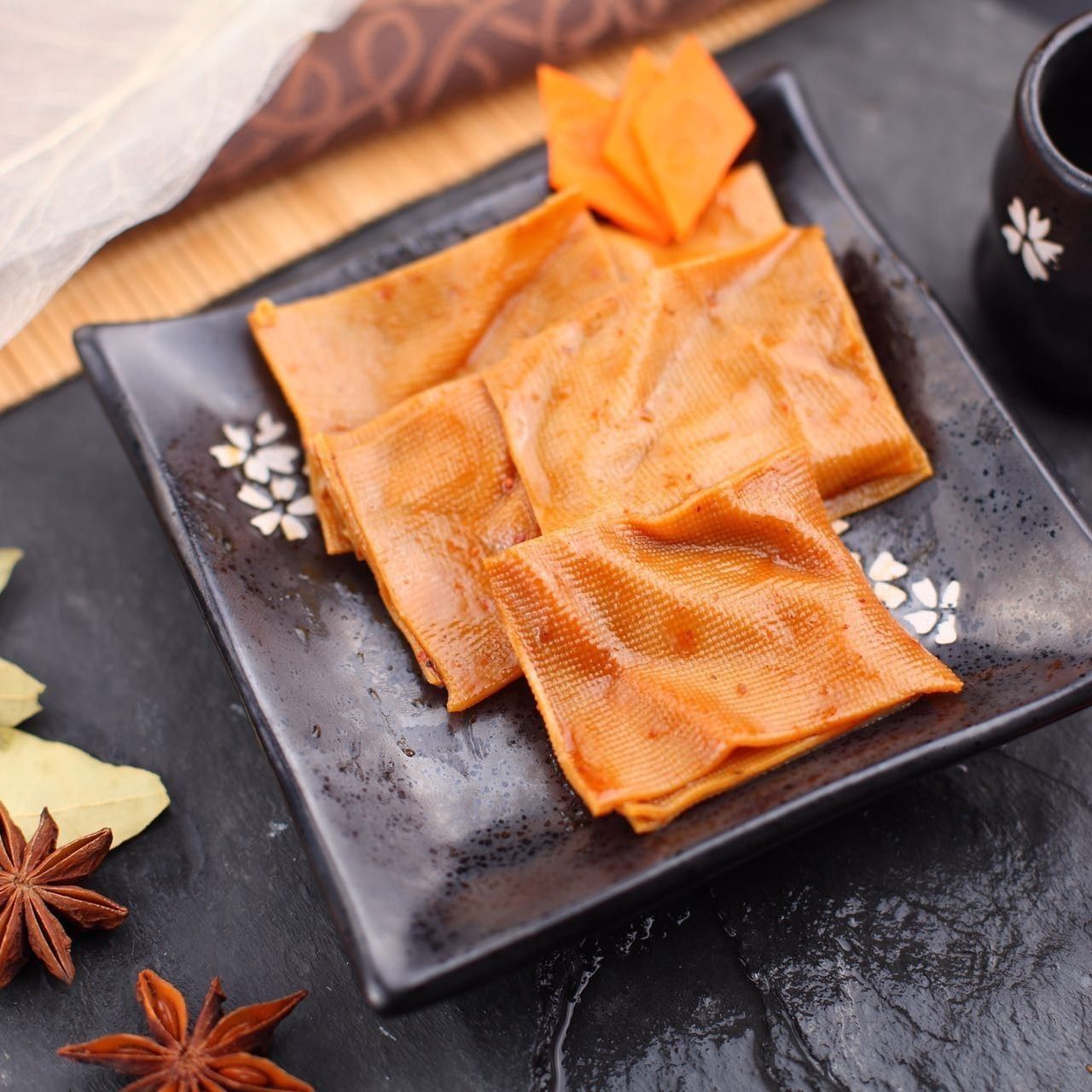 Orange pastry squares on a black plate with star anise and a cup on a dark table