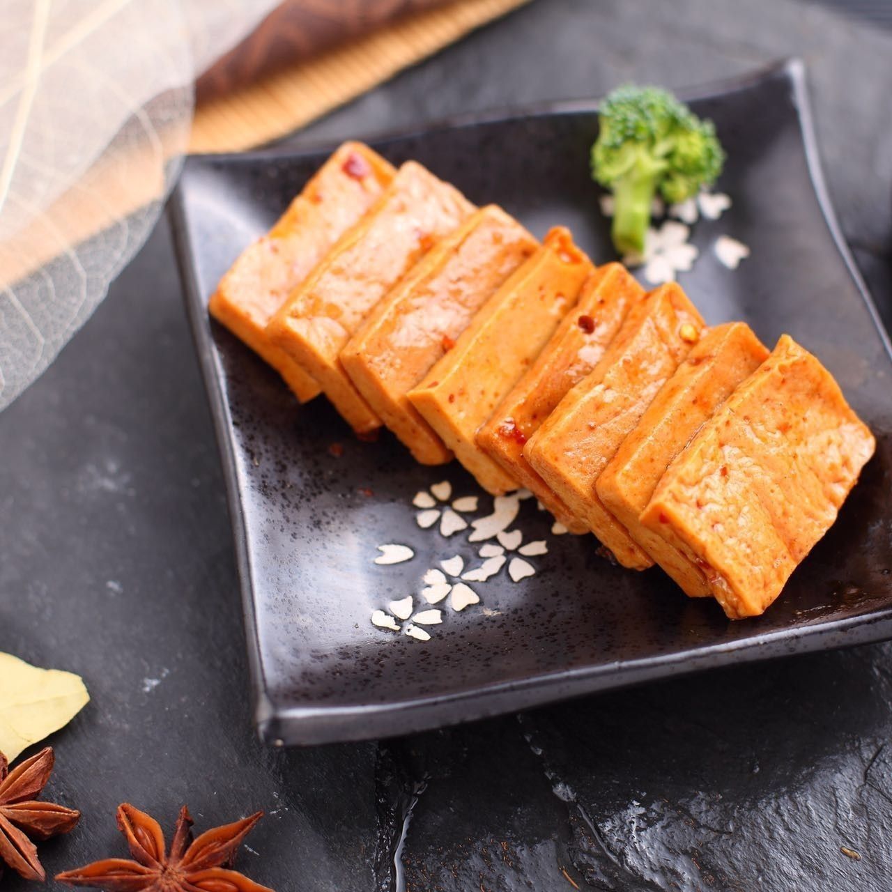 Sliced orange glazed tofu on a black plate, garnished with sesame seeds and broccoli.