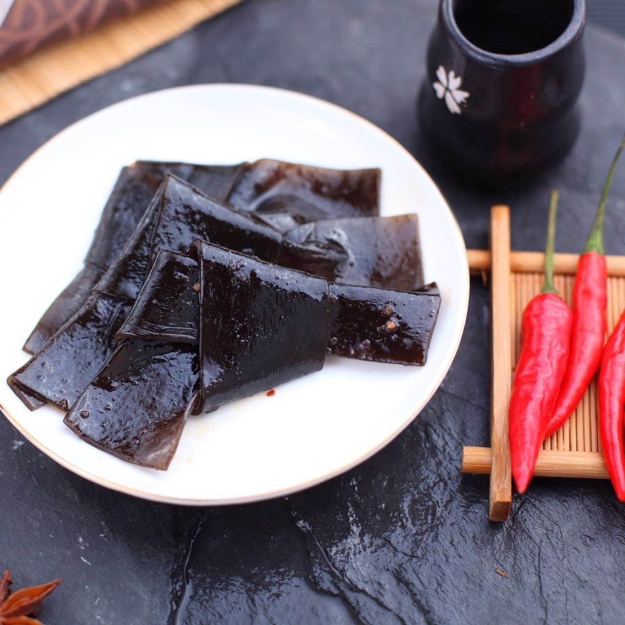 Black gelatin dessert slices on a white plate with red chilies and a dark drink on a slate table