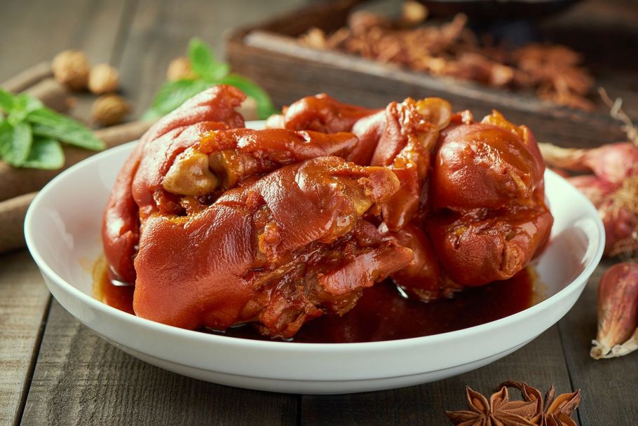 Braised pork knuckle in rich red sauce on a white plate, with star anise and herbs nearby