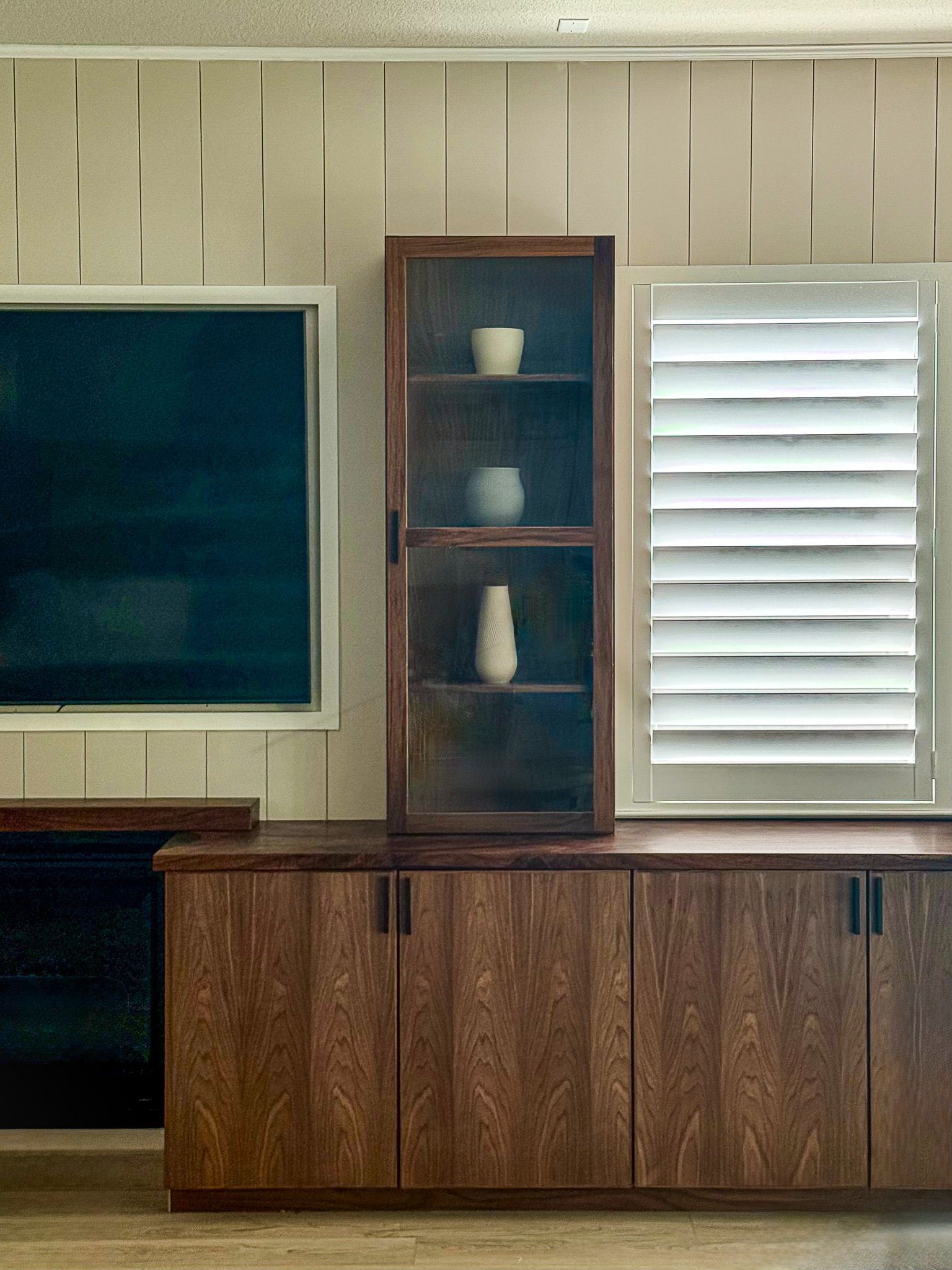 Wooden cabinet with display shelf holding pottery, next to a window with shutters. A dark screen is on the left.