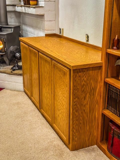 A honey oak storage cabinet with four doors, built into a wall, with a bookshelf to the right.