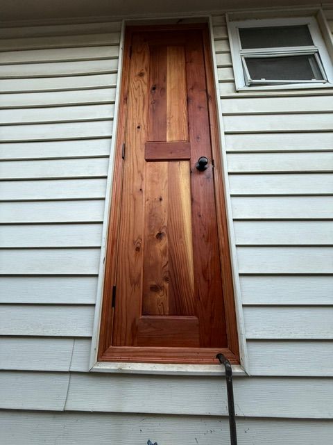 A narrow, wooden redwood door with a crossbar sits on a white-sided building. A small window is to the right.