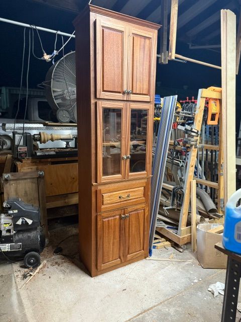 Tall, solid wood cabinet in workshop, brown finish with glass-doored center section, drawer, and top doors.