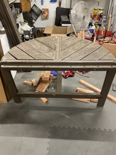 Brown, semi-circular table in a workshop setting, with wood pieces and tools visible in the background.