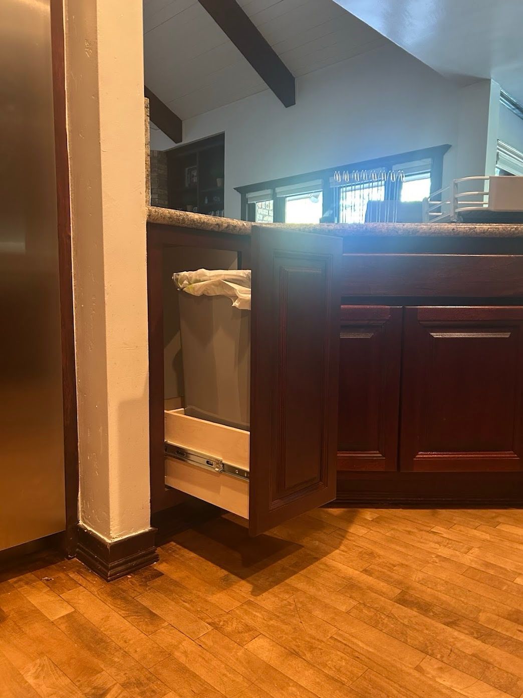 A pull-out trash can in a kitchen with a dark mahogany cabinet. The cabinet is next to a white pillar and wooden floors.