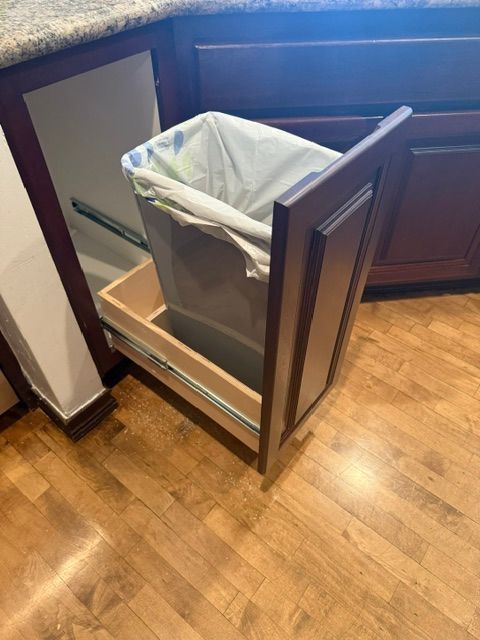 Pull-out trash can cabinet with a partially extended wooden drawer, revealing a trash bag inside.
