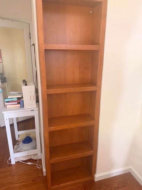 Tall empty wooden bookshelf beside a small white table and mirror in a room with hardwood floor.