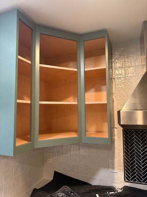 Open corner kitchen cabinets with teal doors and empty wood shelves above a stove backsplash.