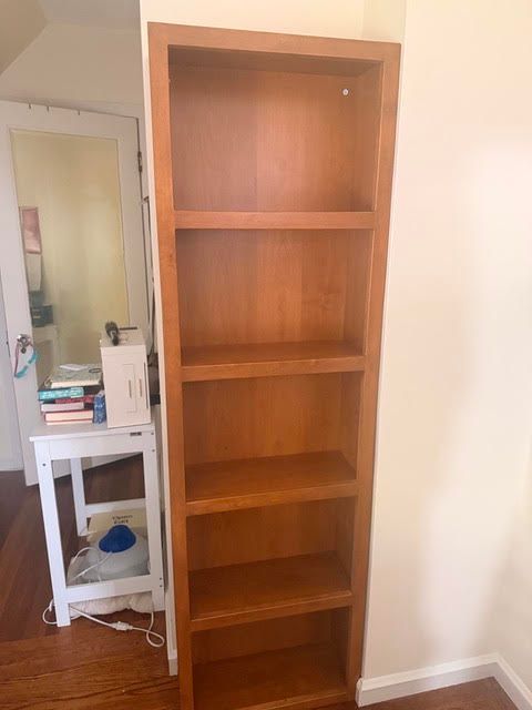 Tall wooden bookshelf beside a desk in a small room, with several empty shelves
