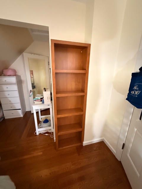 Narrow wooden bookshelf in a mostly empty room with hardwood floors and white walls.