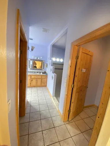Hallway with three wooden-framed doorways; two lead to bathrooms, one open. Light-colored tile floor, blue walls.