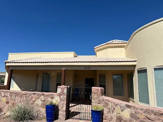 Beige house with stone wall, blue pots, and a black gate under a blue sky.