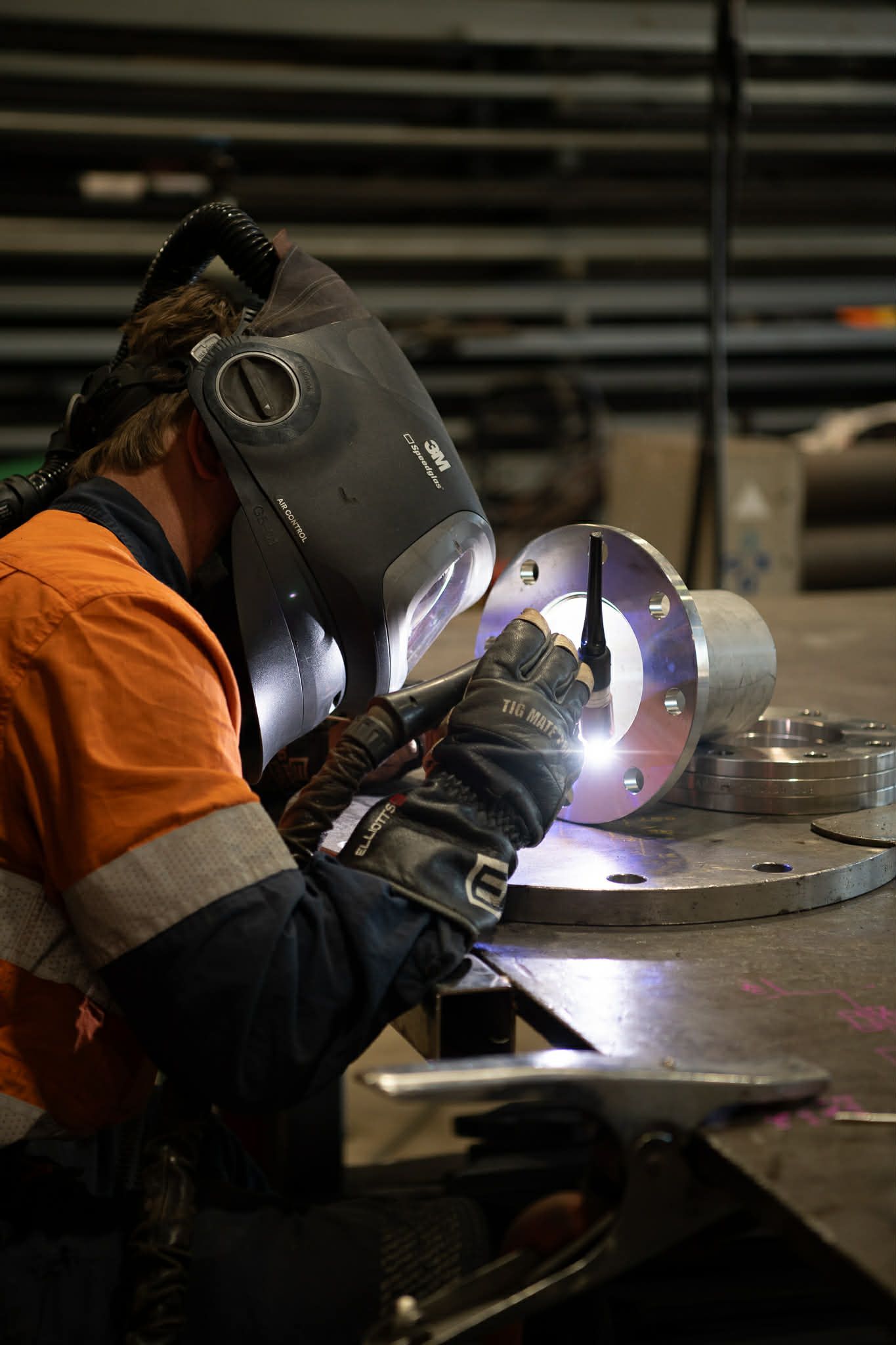 Person welding metal railing on a platform, wearing protective gear, with sparks flying in a factory setting.