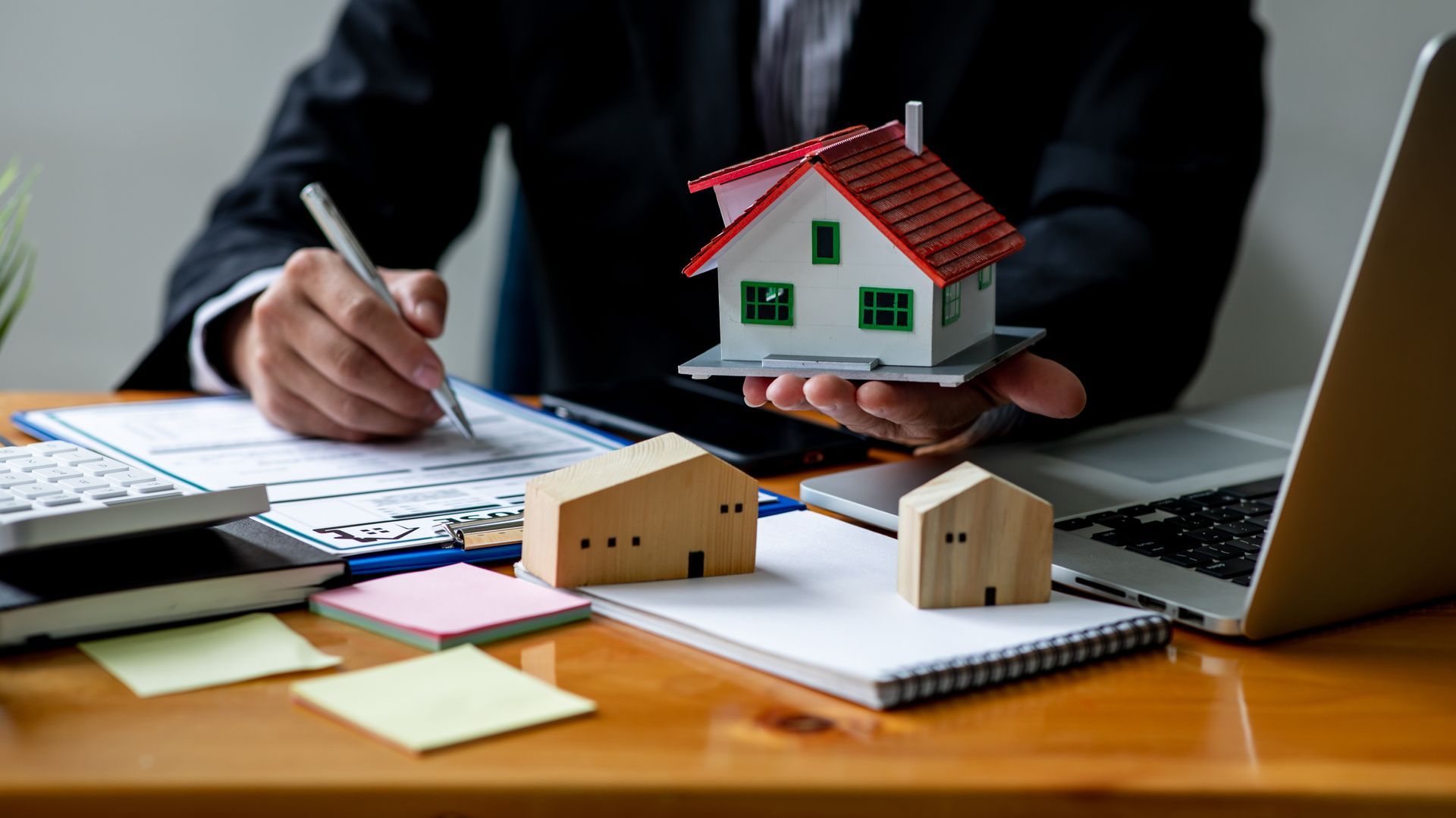A person in a suit holds a small model house while signing home insurance documents at a desk.