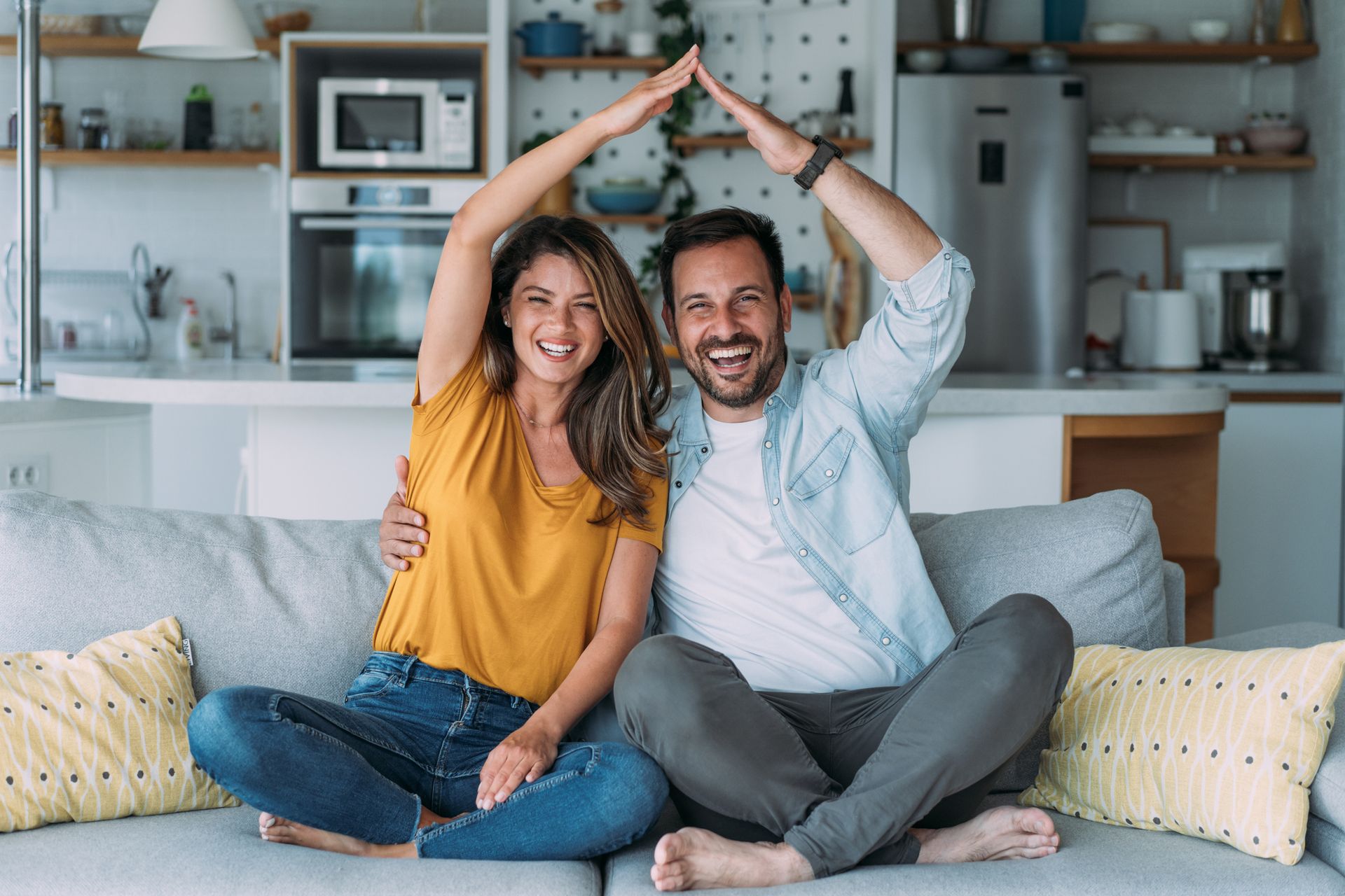A cheerful couple sitting on the sofa, making a house figure with their hands over their heads.