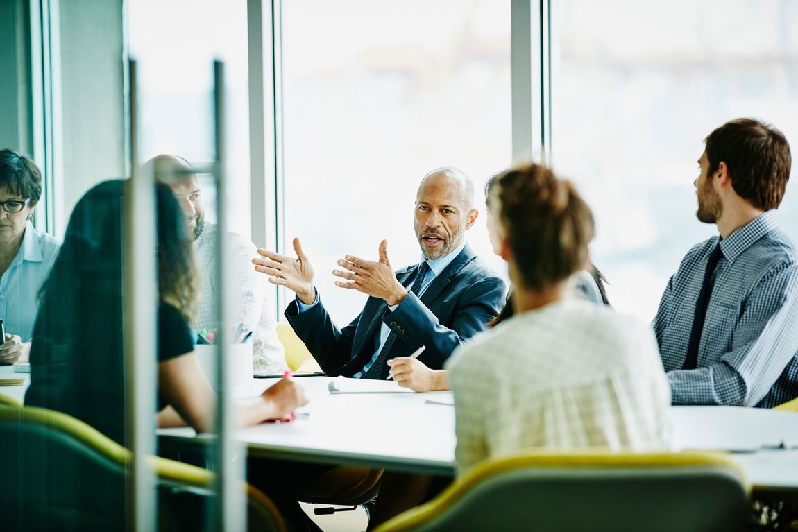 Business meeting in a modern office. A man in a suit gestures as he speaks to colleagues around a table.