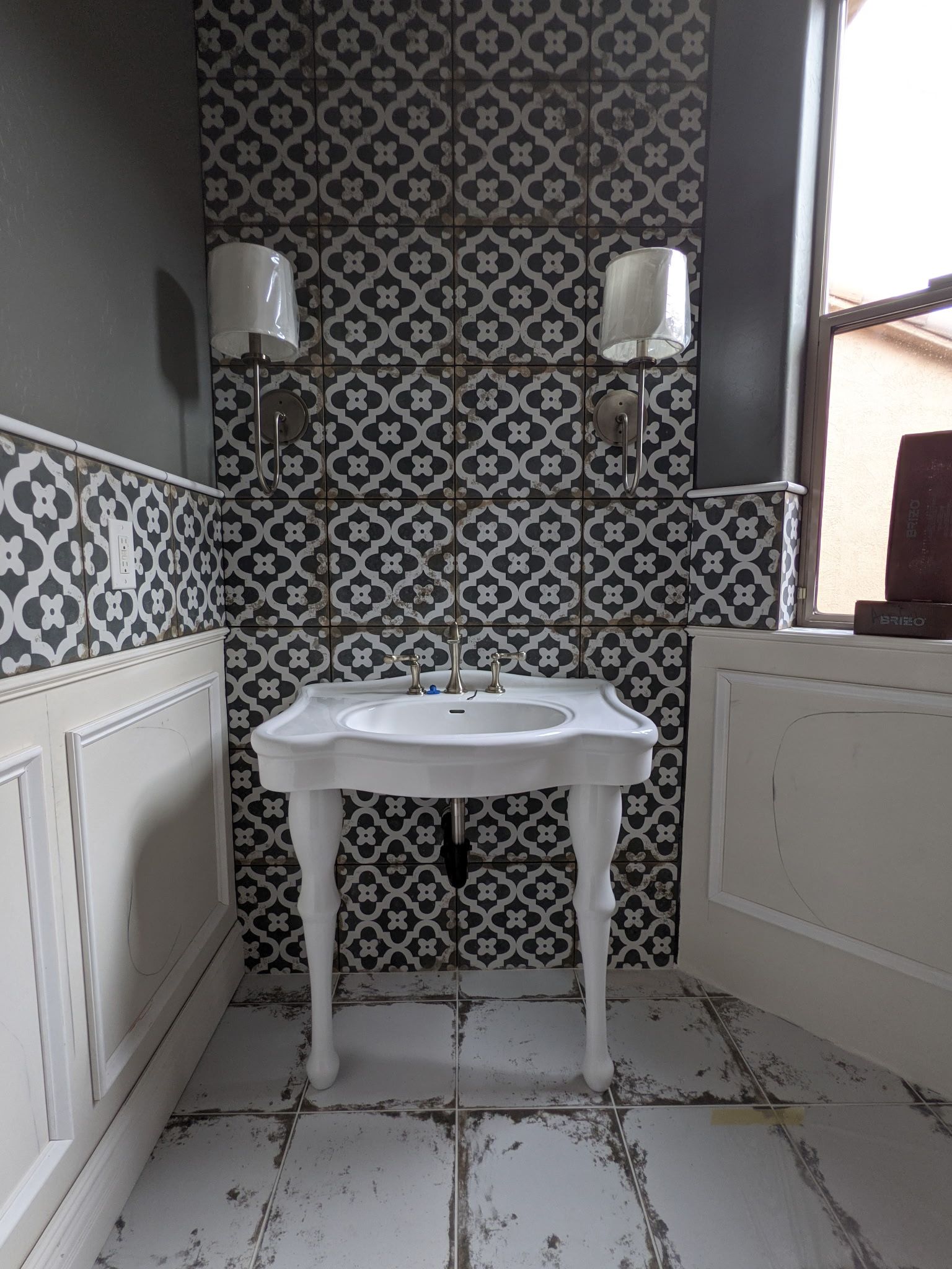 Bathroom with patterned tile wall behind a white pedestal sink and sconce lights.