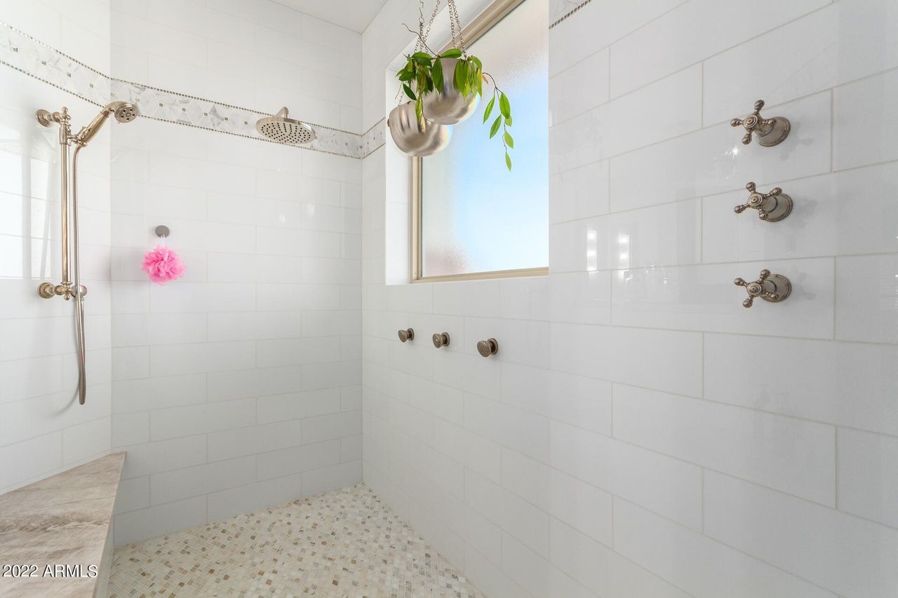 White tiled shower with multiple shower heads, window, and hanging plant.