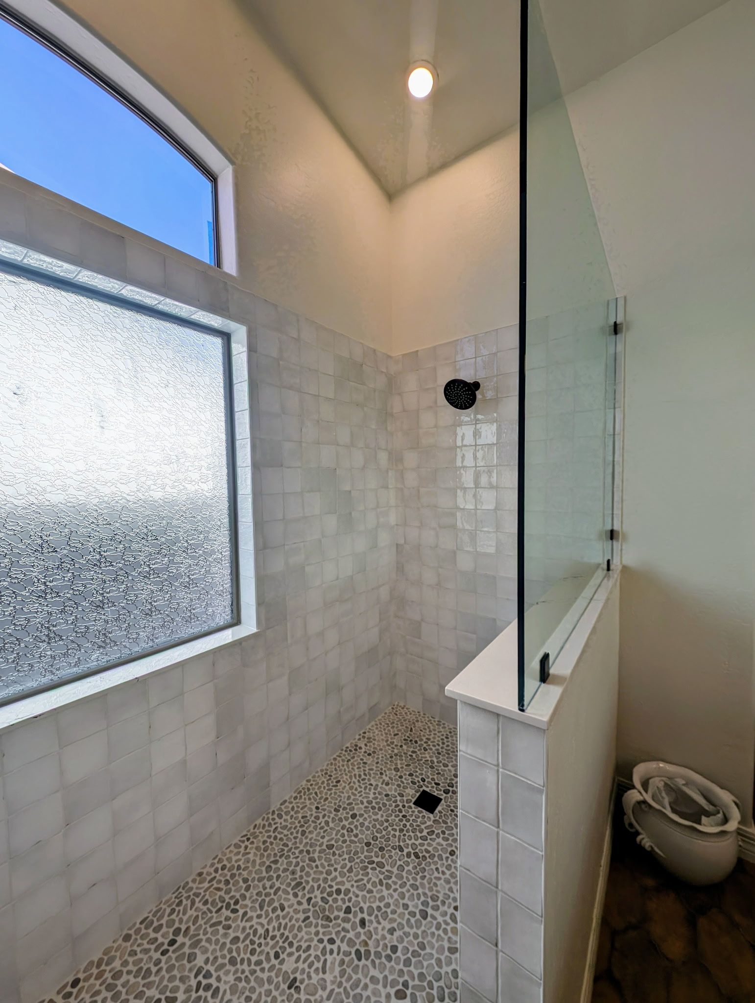 Shower with pebbled floor, tiled walls, glass partition, and small window with textured glass.