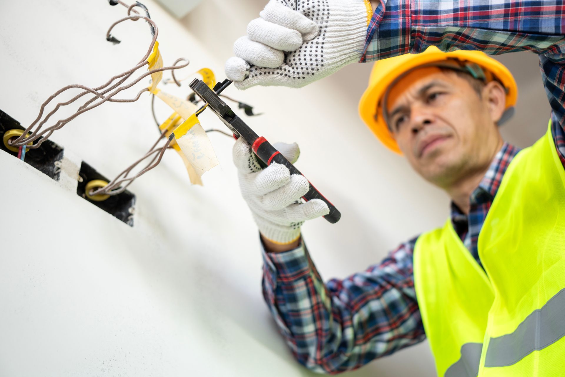 Electrician in yellow hard hat inspecting exposed wiring with a tool in a white wall cavity.