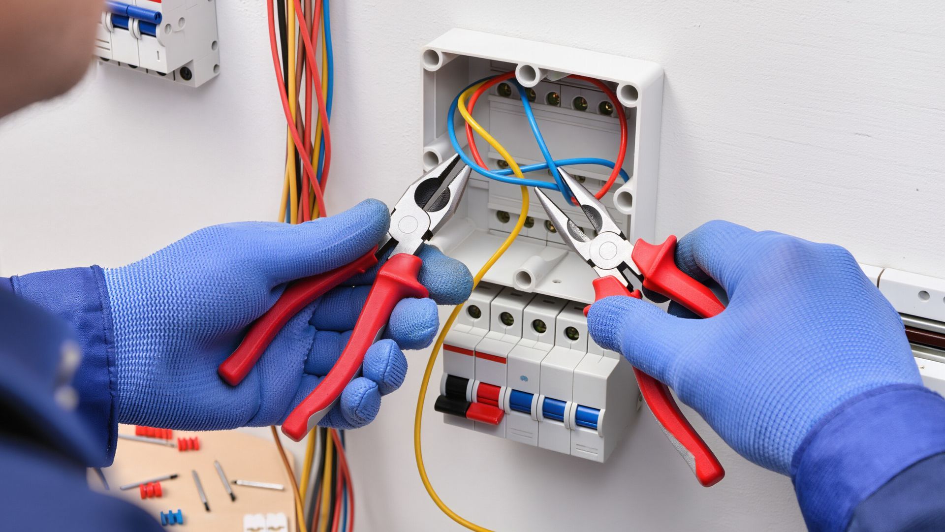 Gloved hands use a screwdriver and pliers to work on exposed wiring.