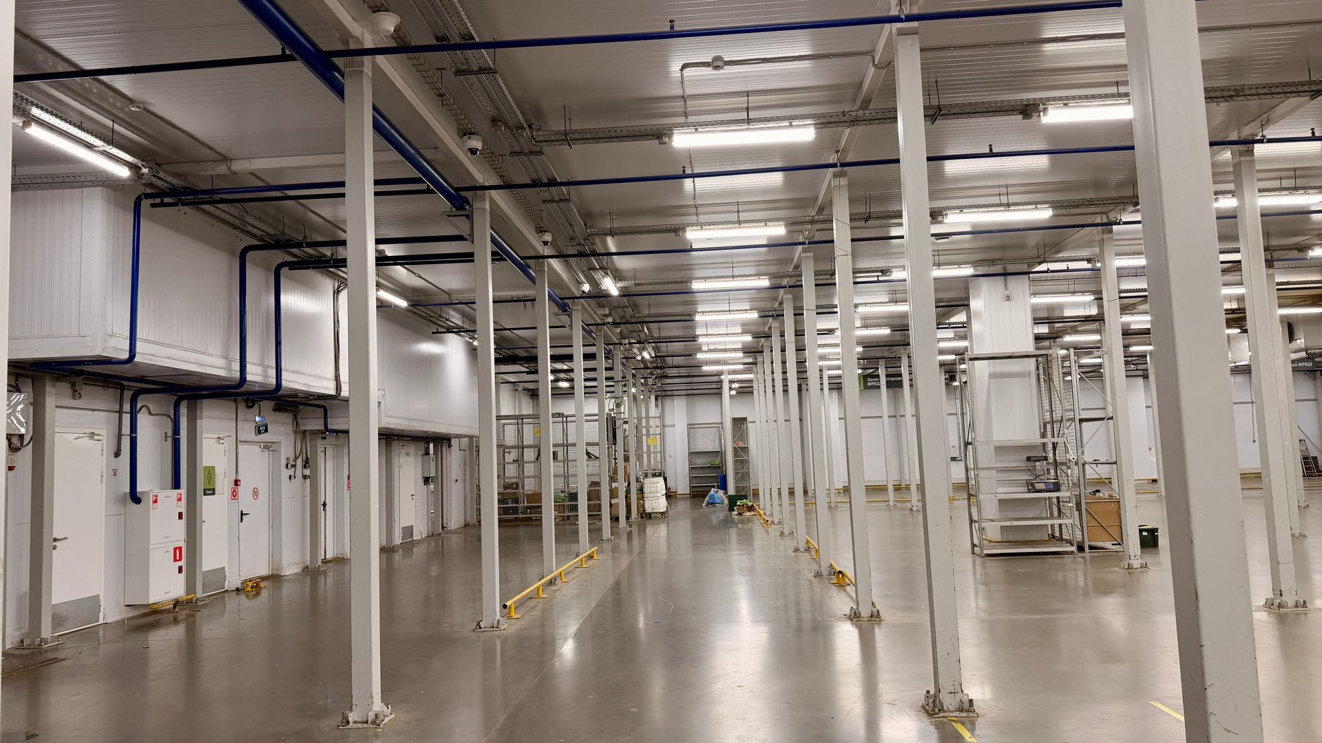 Large industrial warehouse interior with tall white columns, metal shelving, and bright overhead lights.