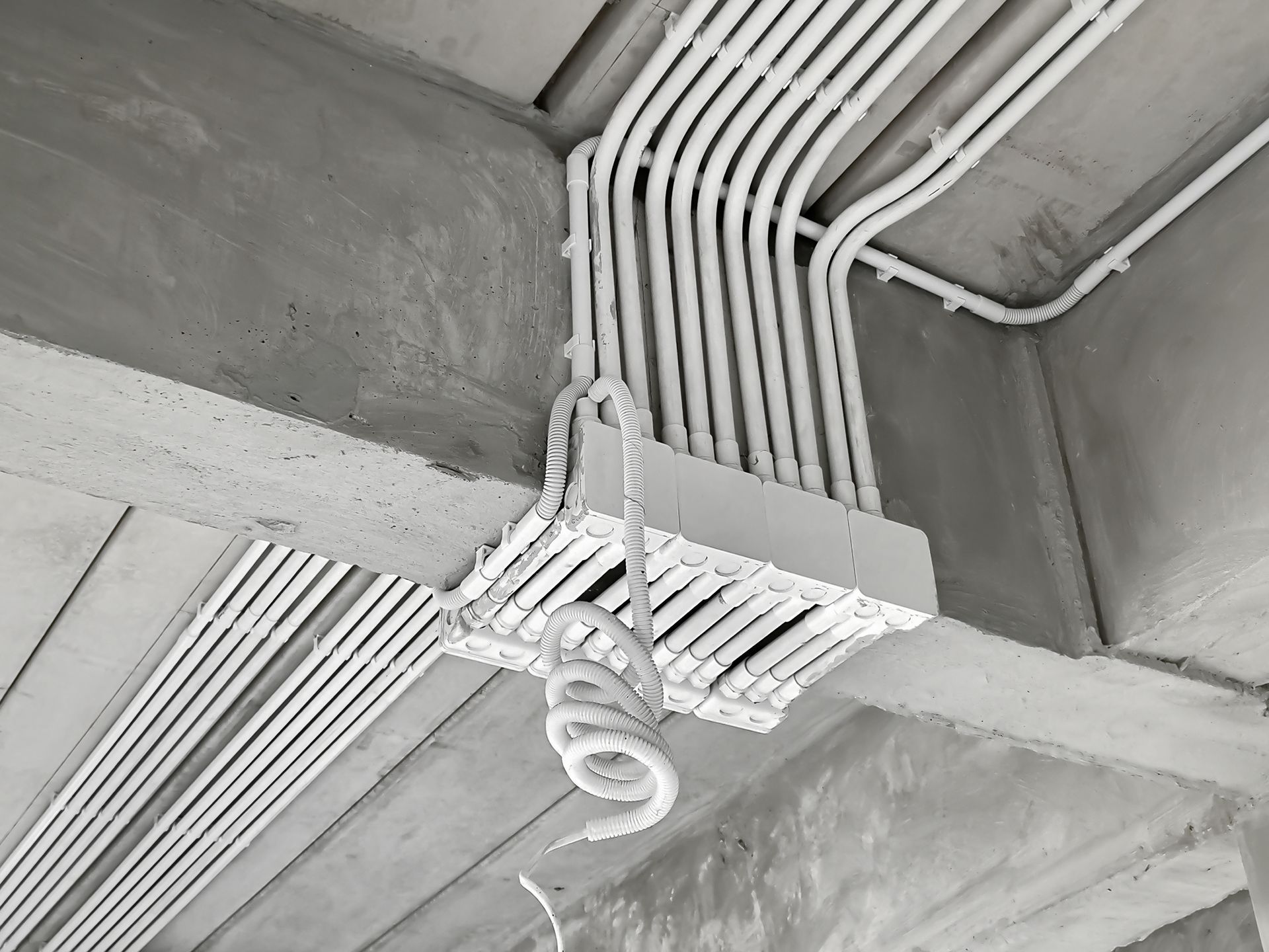Ceiling with bundled white electrical conduits and a hanging coiled cable in a concrete room.