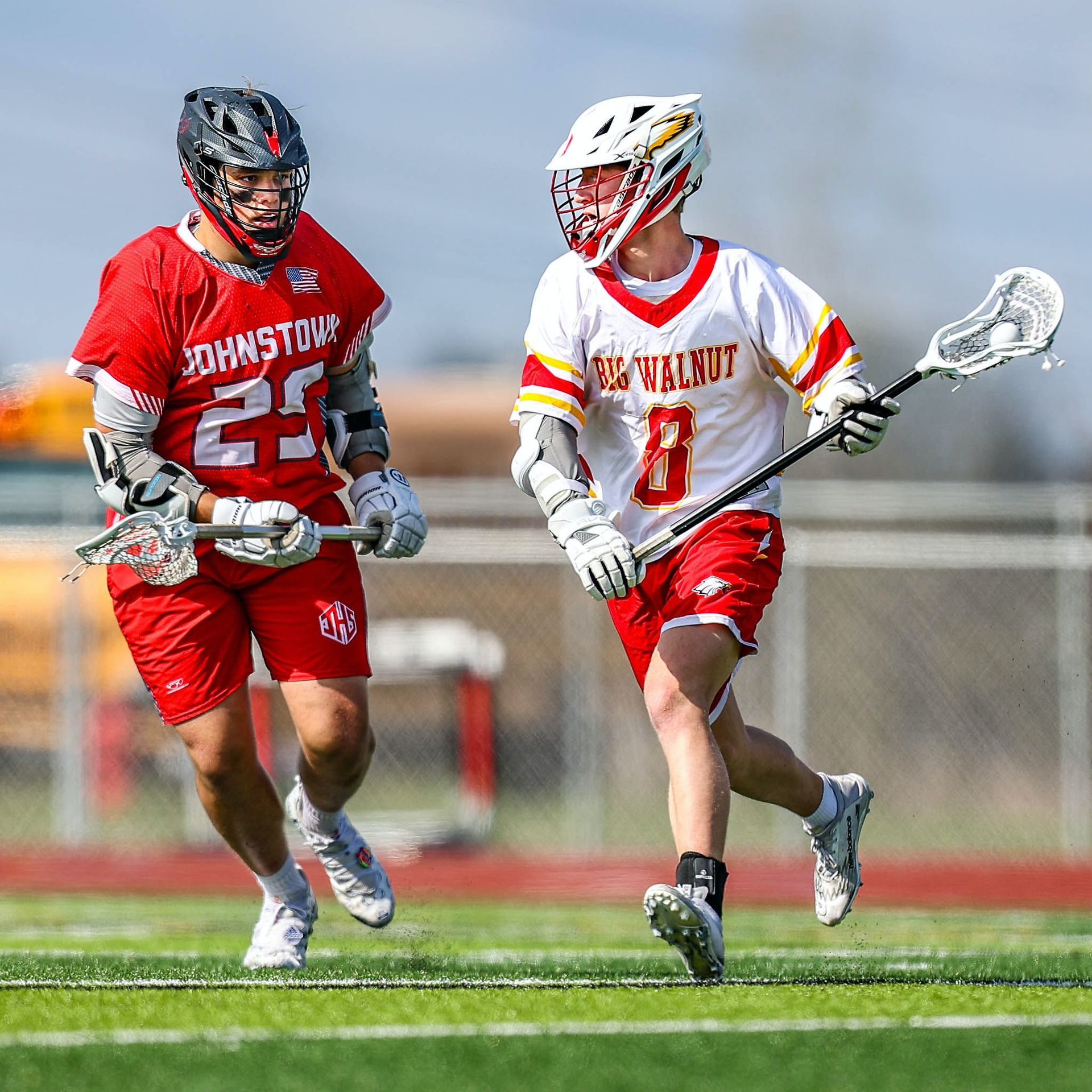 Two lacrosse players in red uniforms running on a field, one cradling the ball with a stick