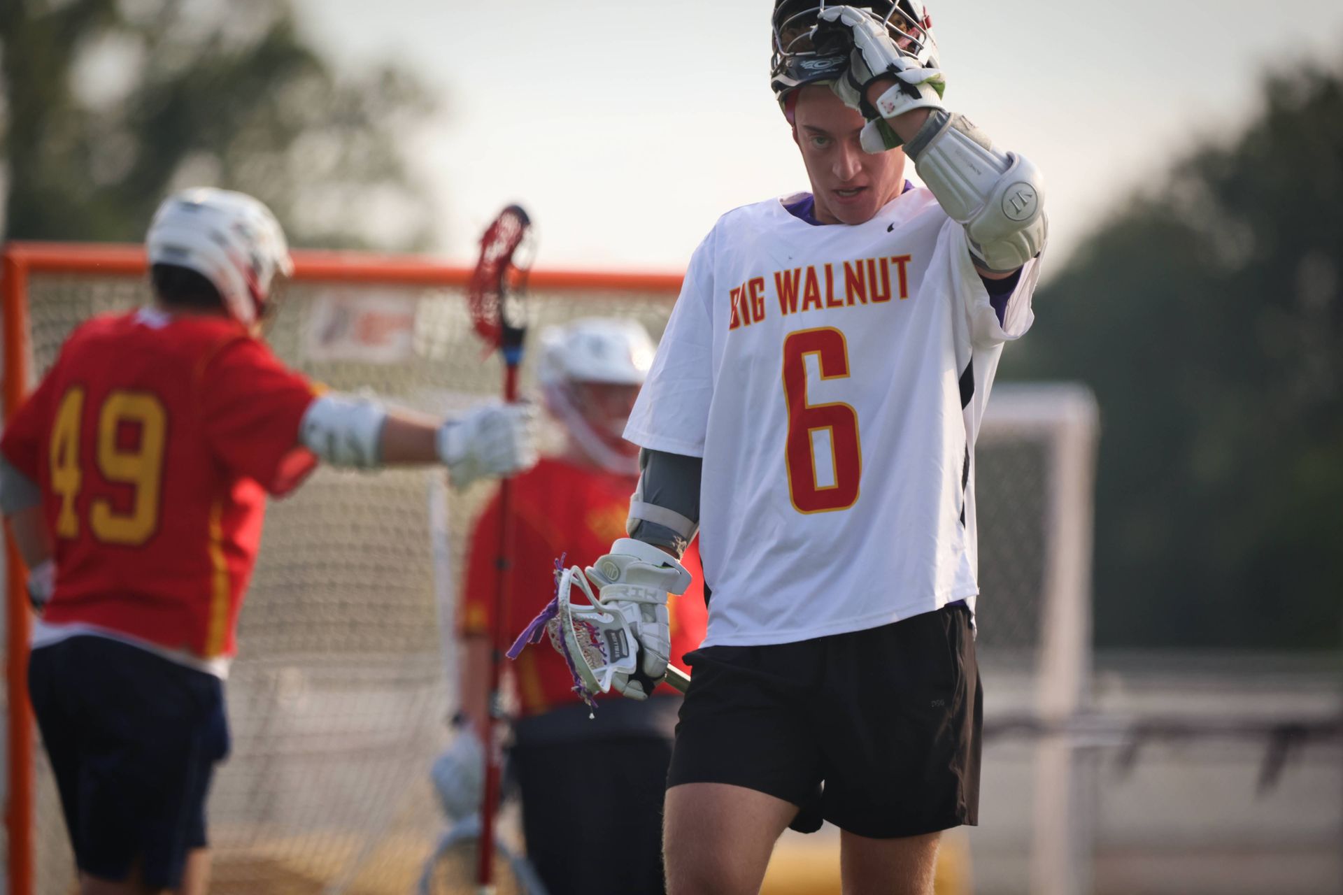 Lacrosse player in white and orange jersey stands on field near goal, adjusting helmet during practice