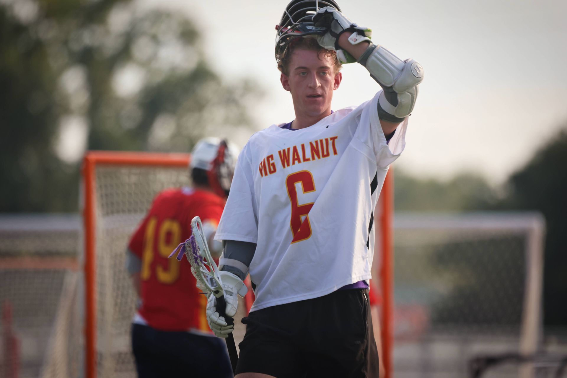 Lacrosse player in white jersey with red number 6 adjusts his headgear on the field near the goal.