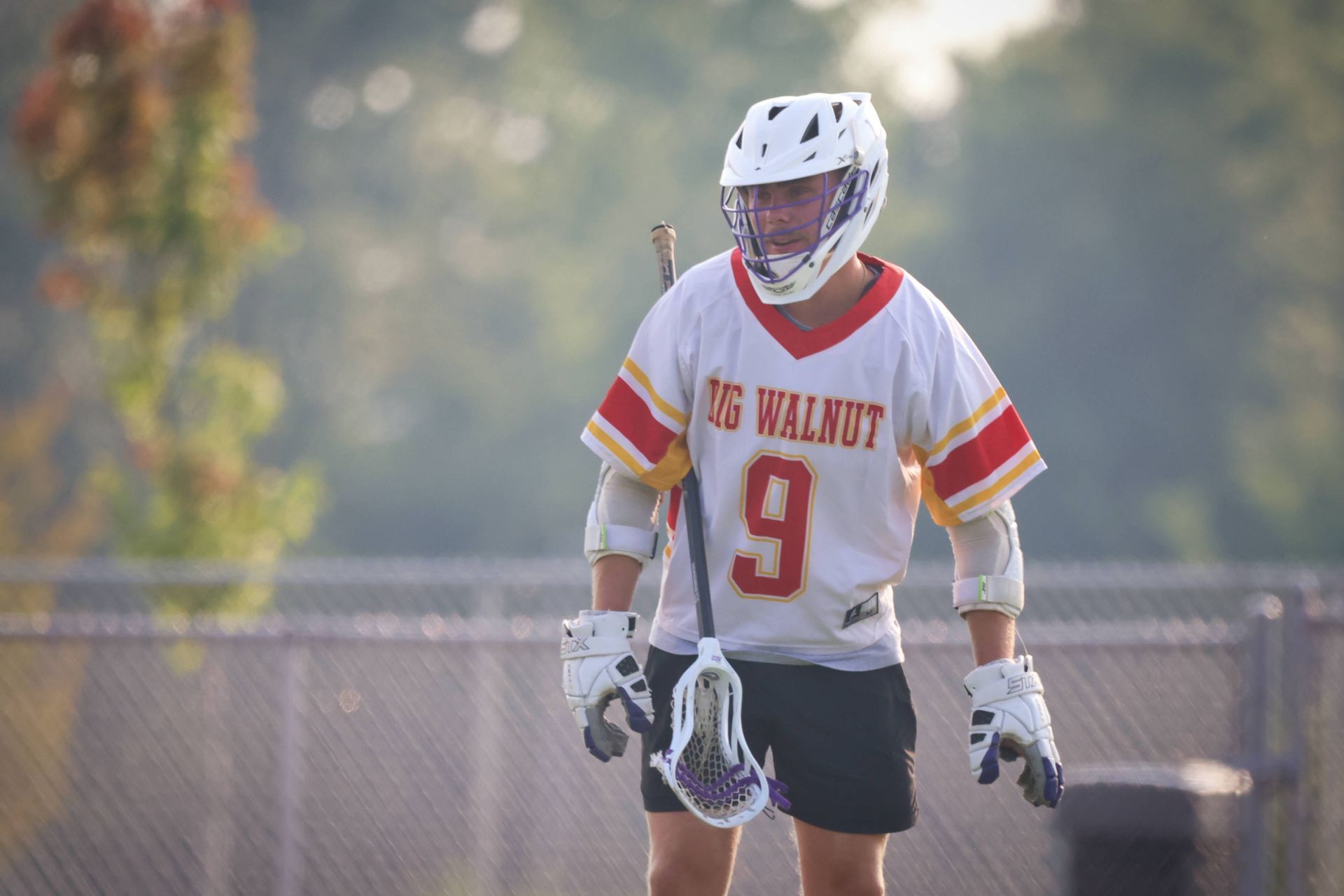 Lacrosse player in white and red uniform with number 9 stands on a field holding a stick.