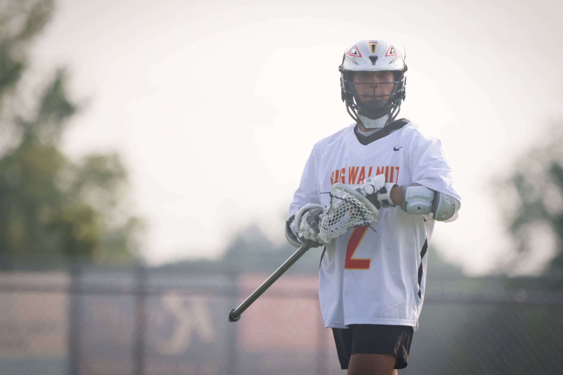 Lacrosse player in white jersey holding a stick on an outdoor field at sunset.