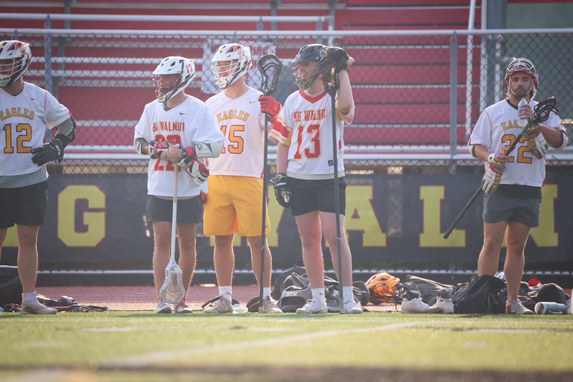 Lacrosse players in white and red uniforms standing along the sideline on a sunny field.