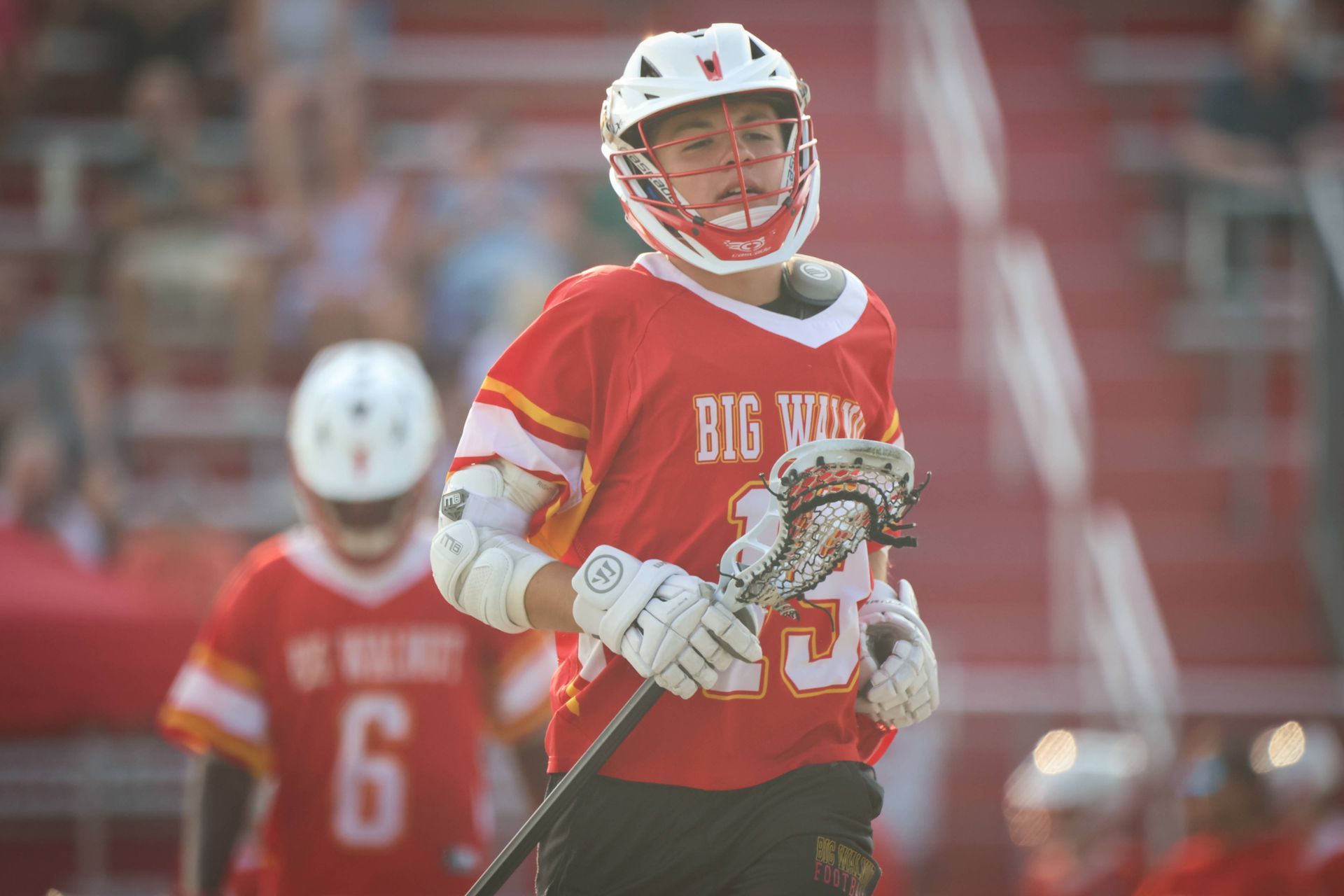 Lacrosse player in red Big Yarmouth jersey holding a stick on the field, with a blurred teammate behind.