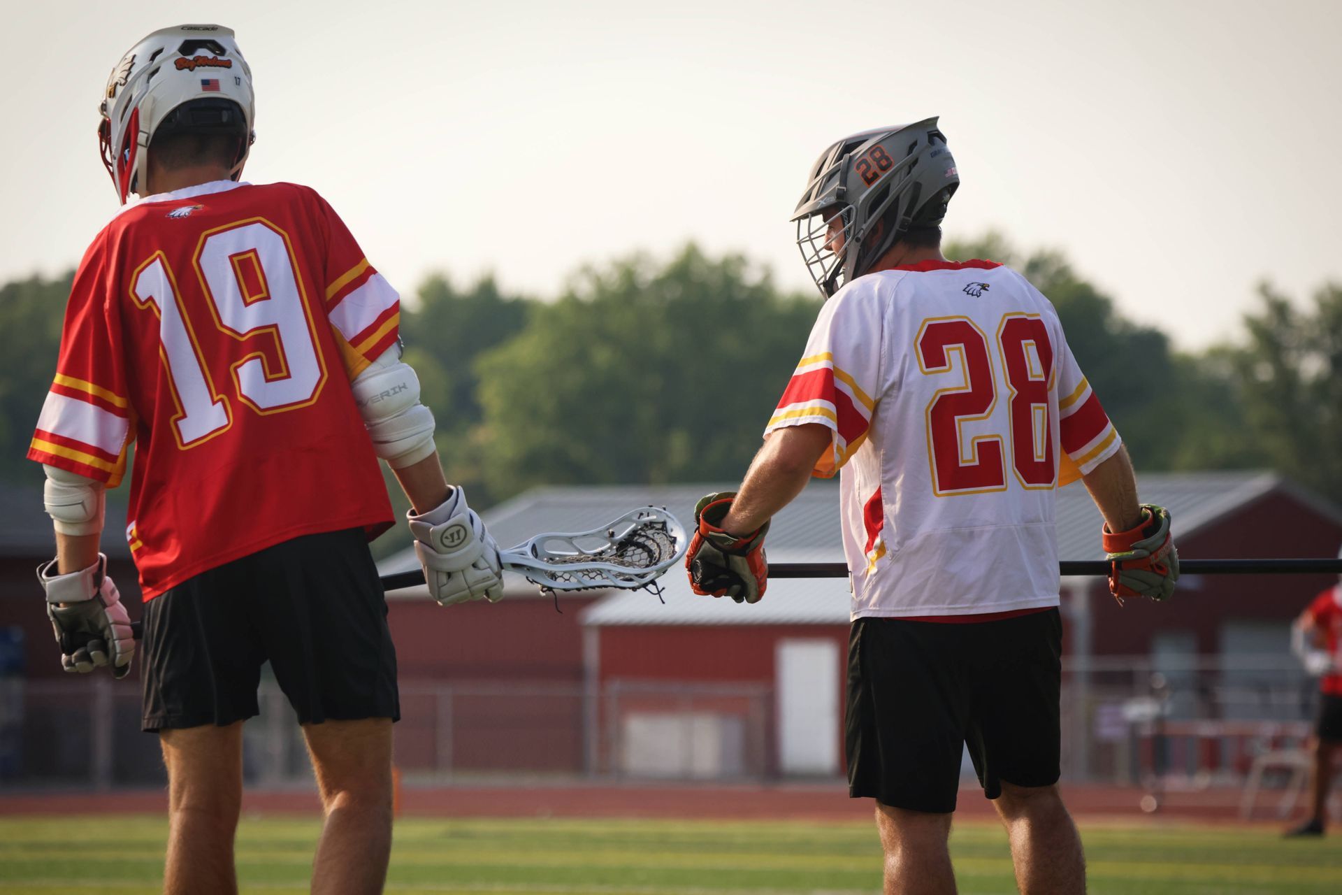 Two lacrosse players in red and white jerseys standing on a field at sunset