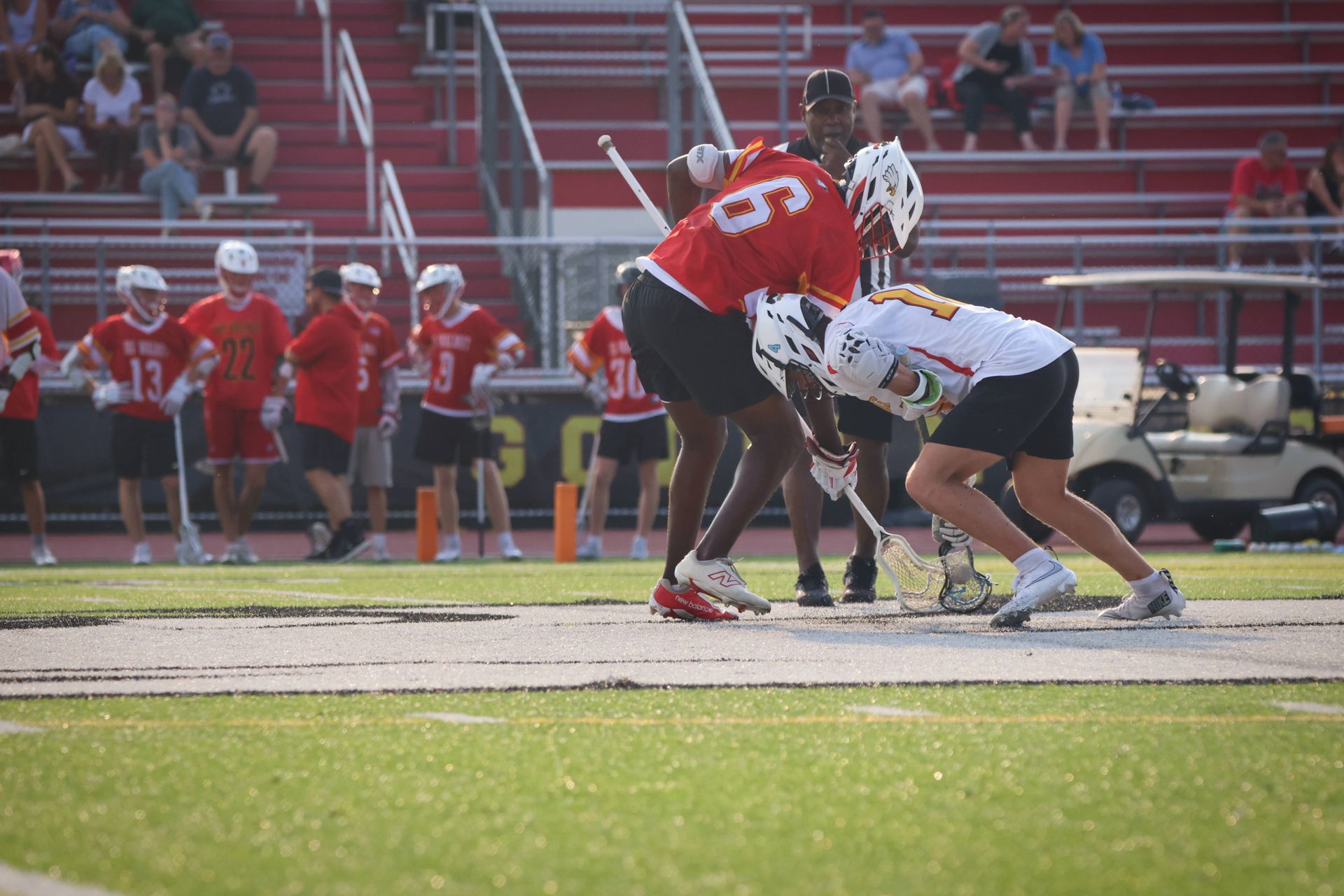 Two lacrosse players in red and white battle for the ball on a field with spectators in the stands.