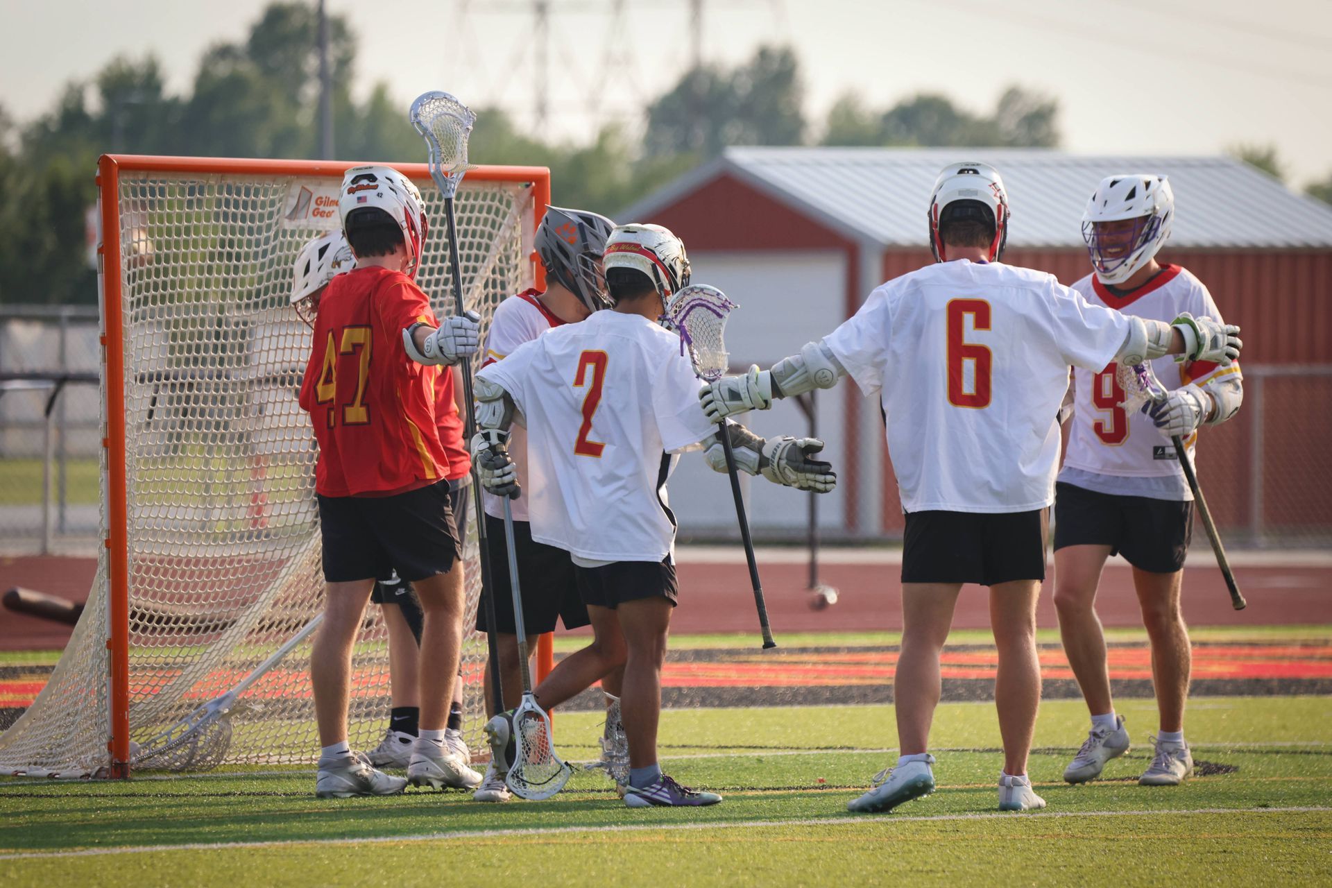 Lacrosse players in white jerseys gather around the goal on a field during a game.