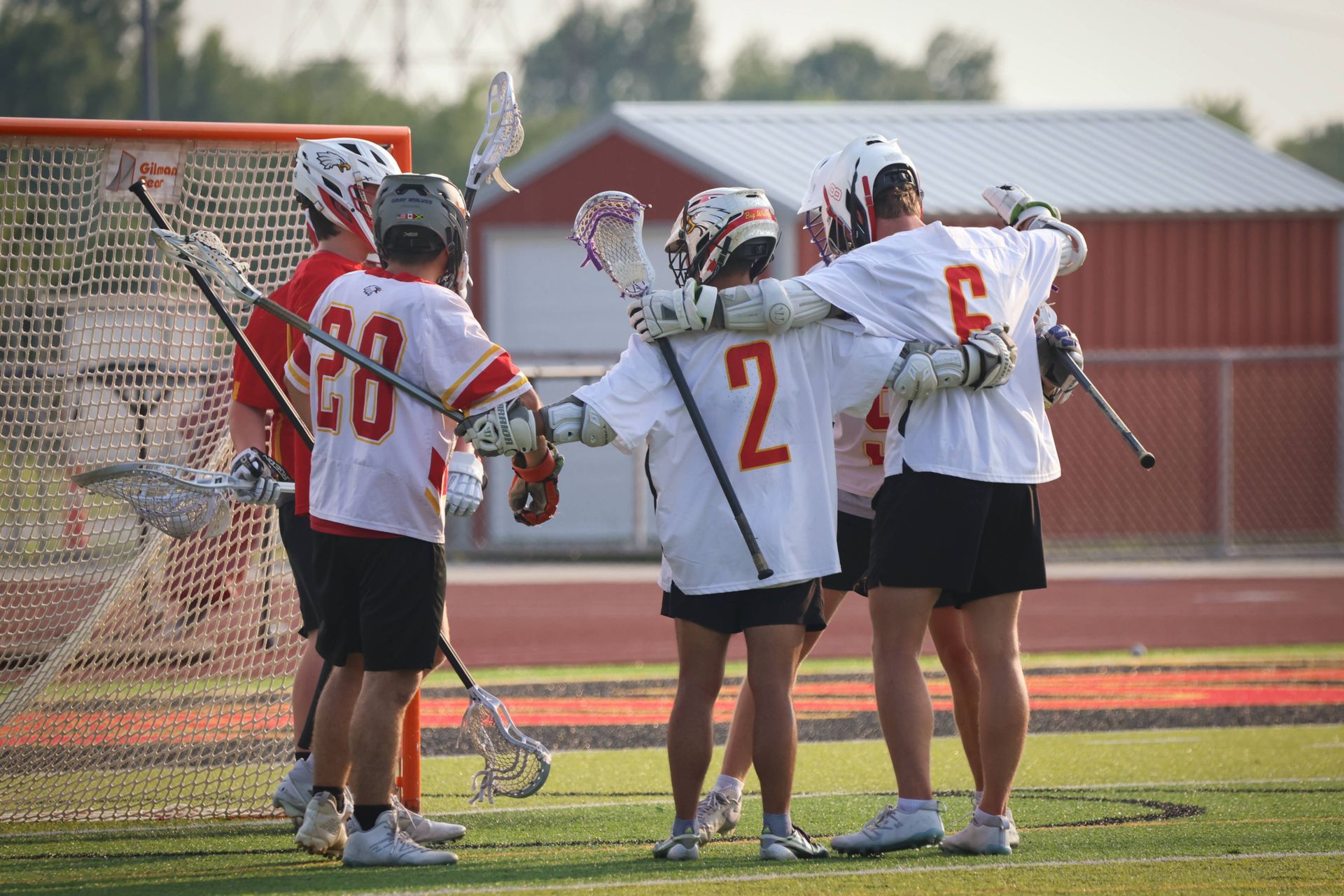 Lacrosse players in white and red jerseys huddle by the goal on a field at sunset.