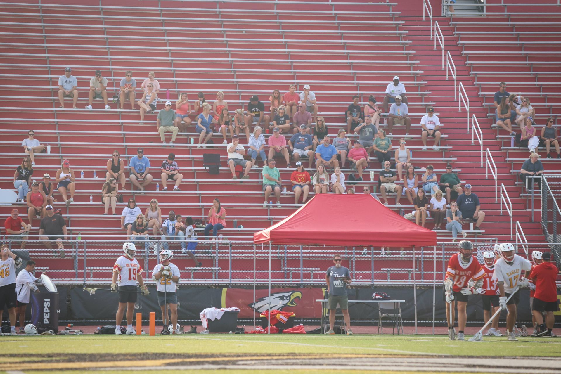 Lacrosse stadium bleachers with scattered spectators and a red tent on the sideline