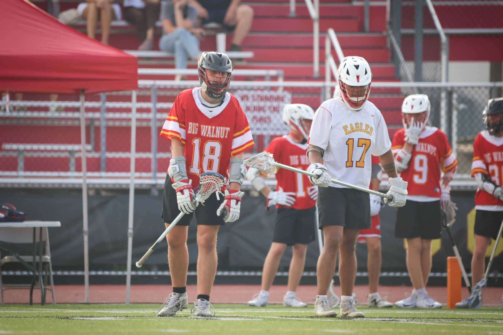 Two lacrosse players in red and white uniforms stand on a field near red stadium bleachers.