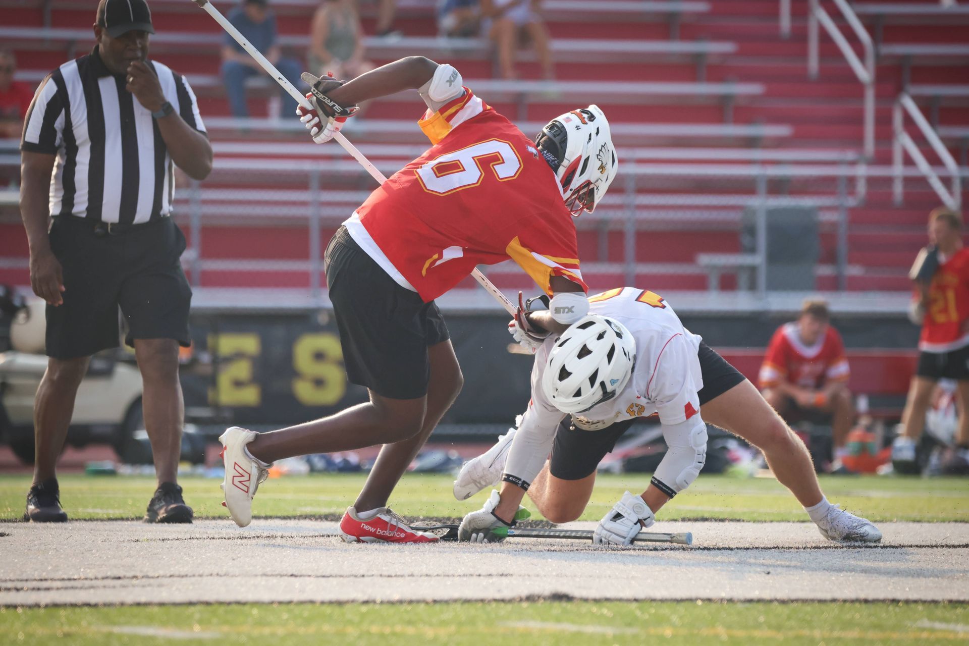 Two lacrosse players battle for the ball on a field, one in red standing over a crouched player in white.