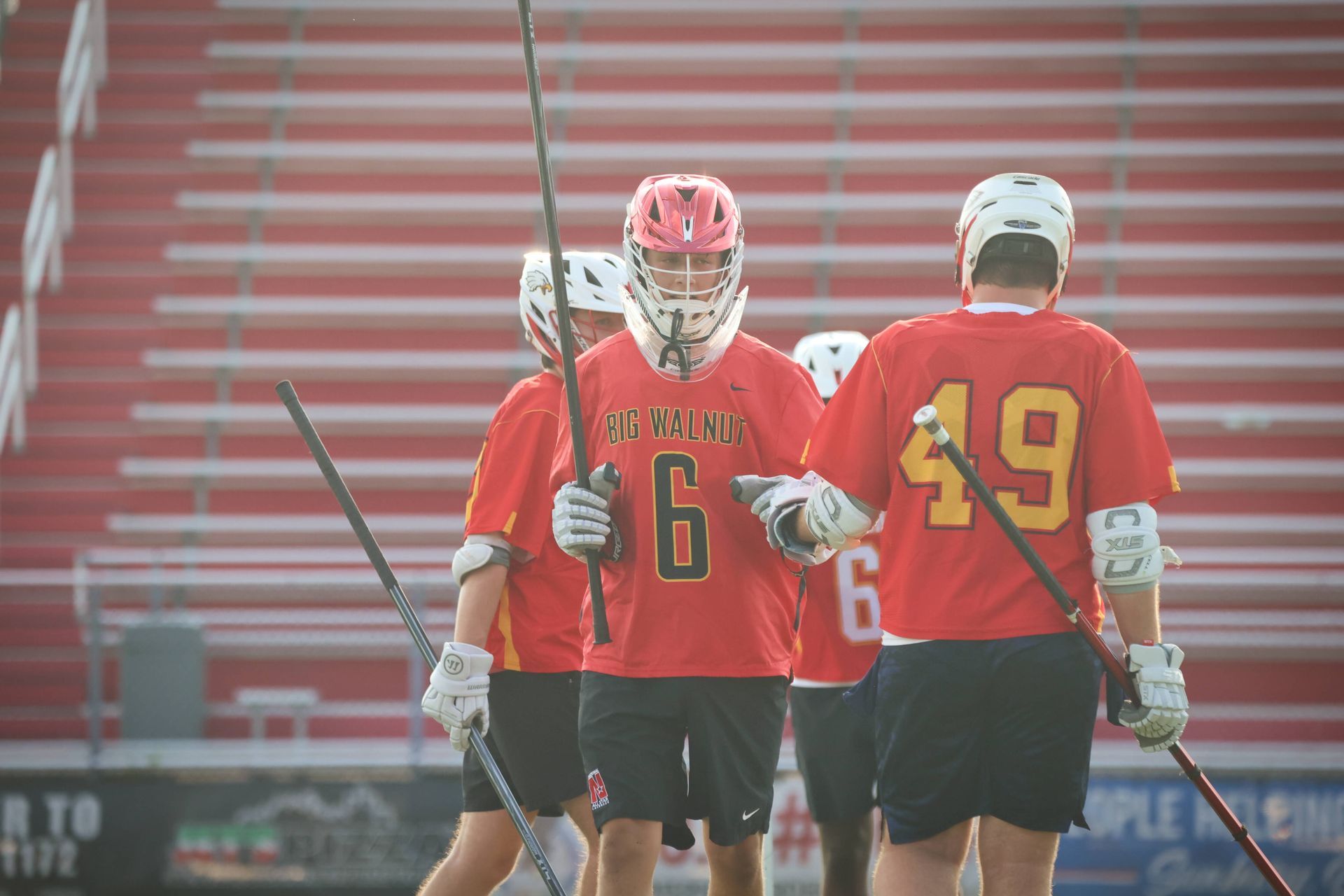 Three lacrosse players in red jerseys huddle on the field with red bleachers behind them.