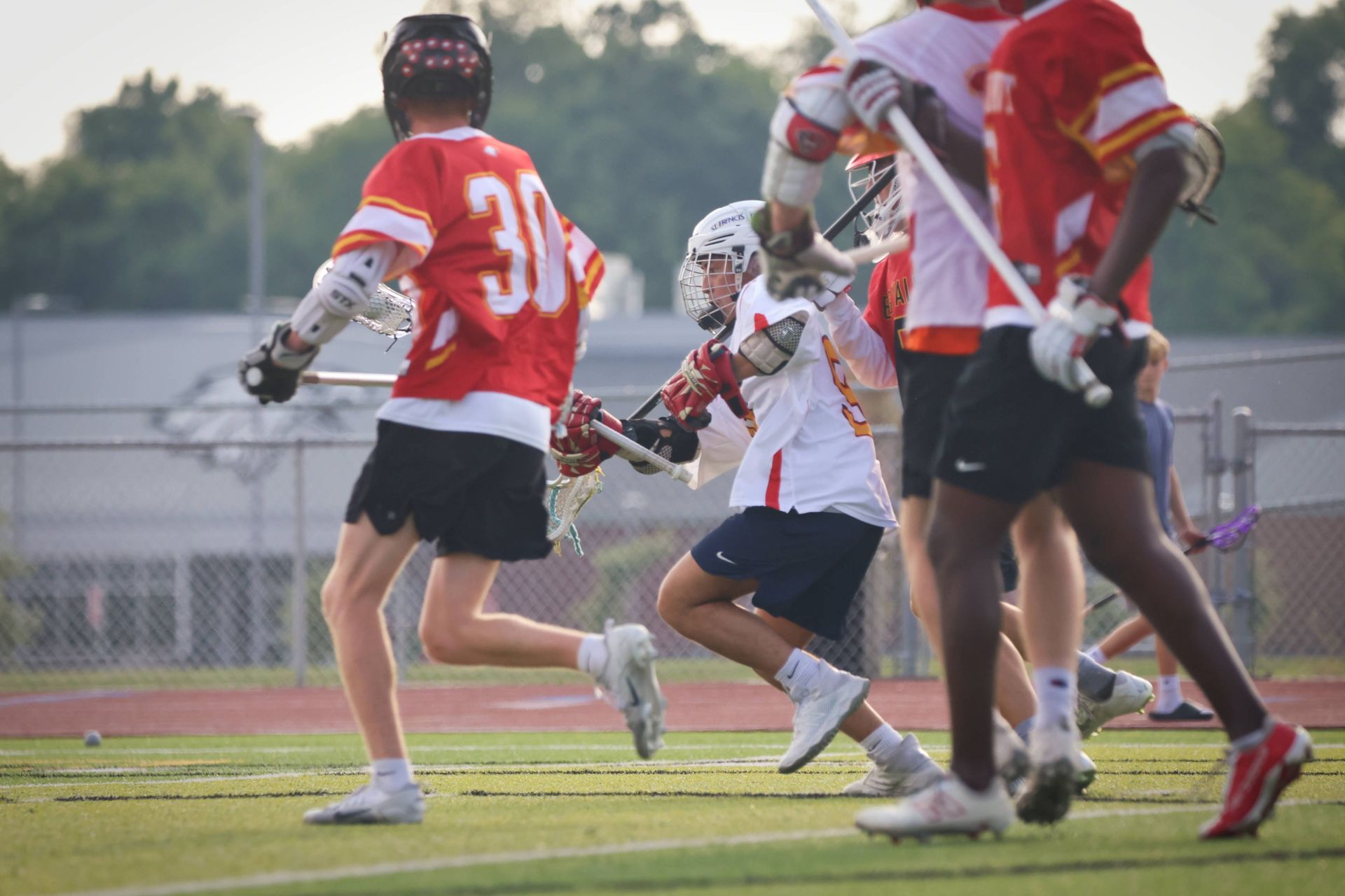 Lacrosse players in red and white uniforms running on a field during a game.