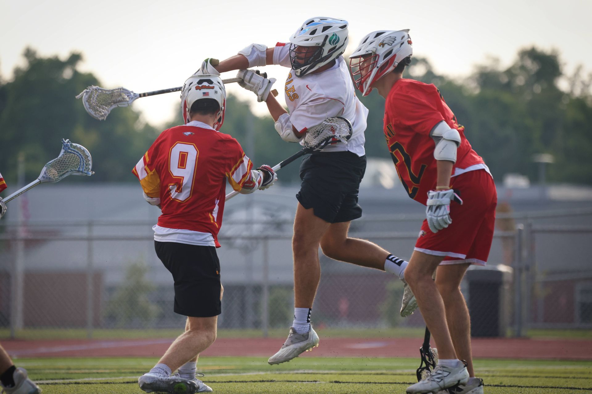 Three lacrosse players in helmets jostle for the ball on a field near dusk