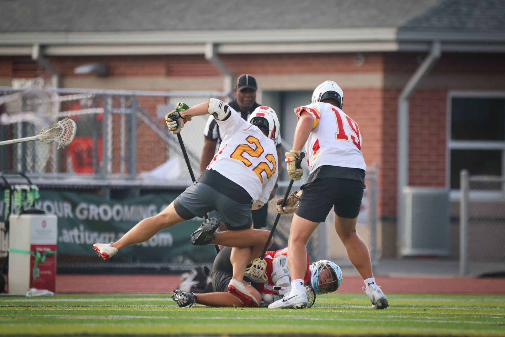 Lacrosse players in white and yellow jerseys battle for the ball near the goal on a field.