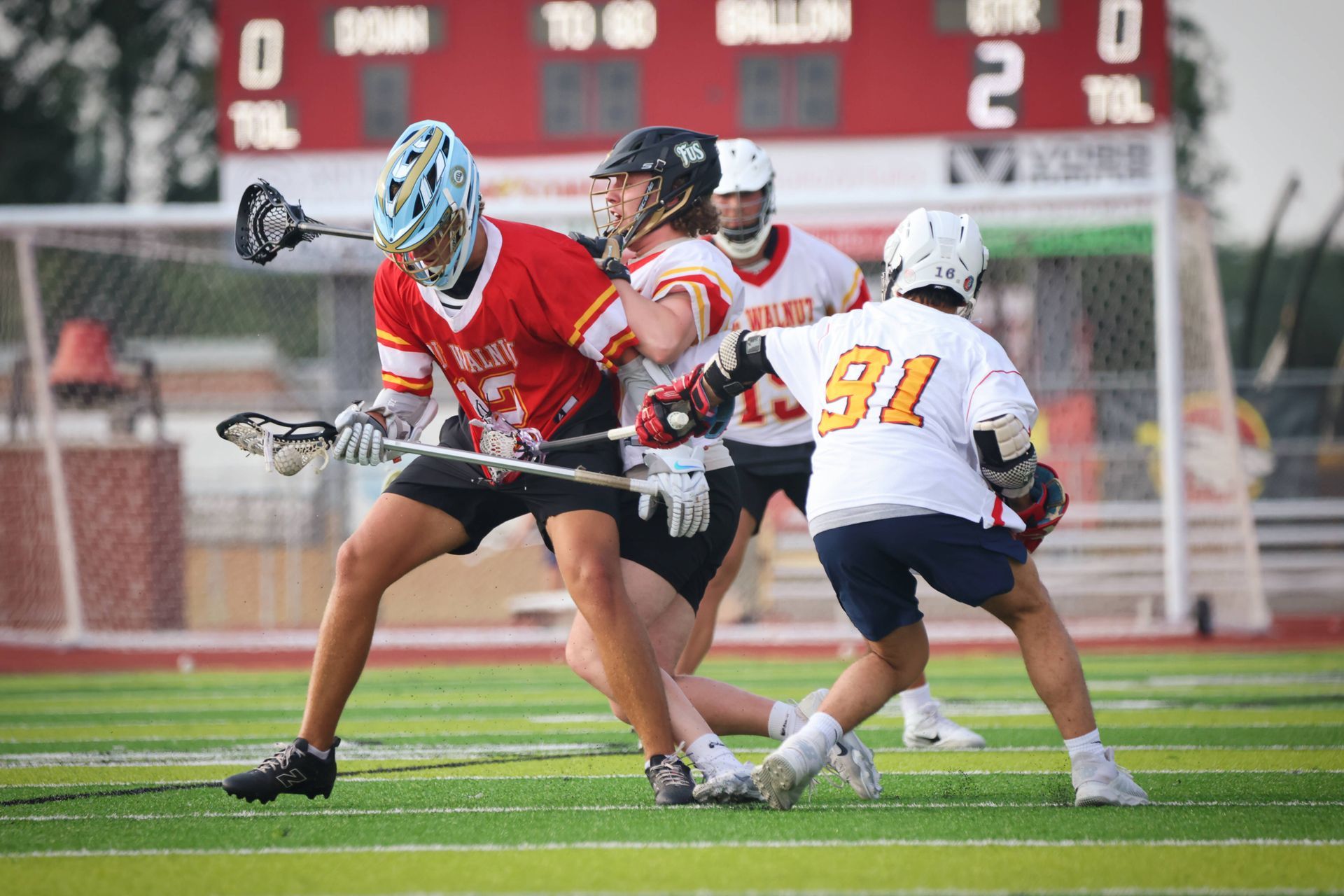 Three lacrosse players battle for the ball on a field, with a scoreboard visible in the background.